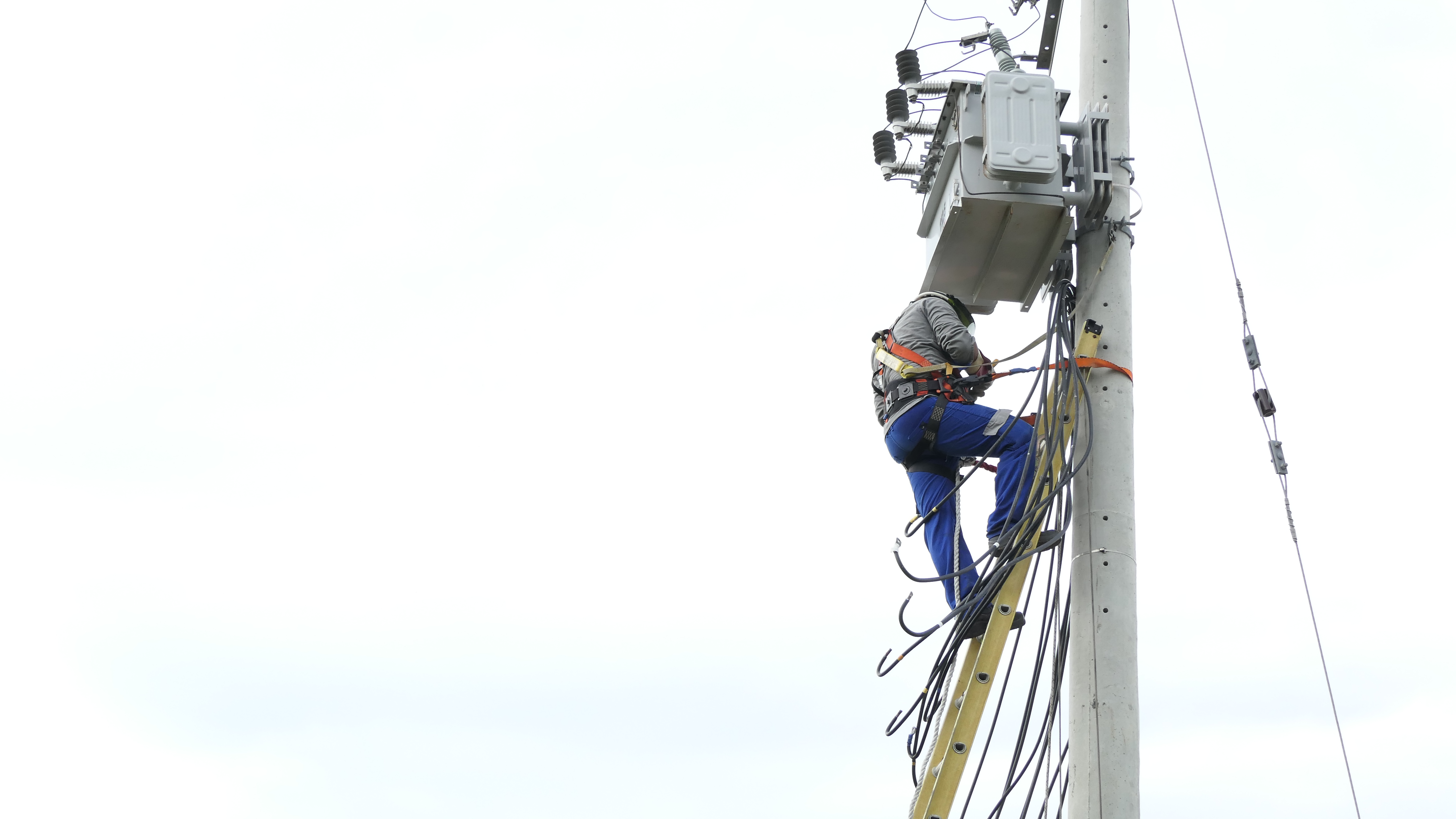 Un trabajador en un poste, ajustando una caja eléctrica, con equipo de seguridad visible en su vestimenta.