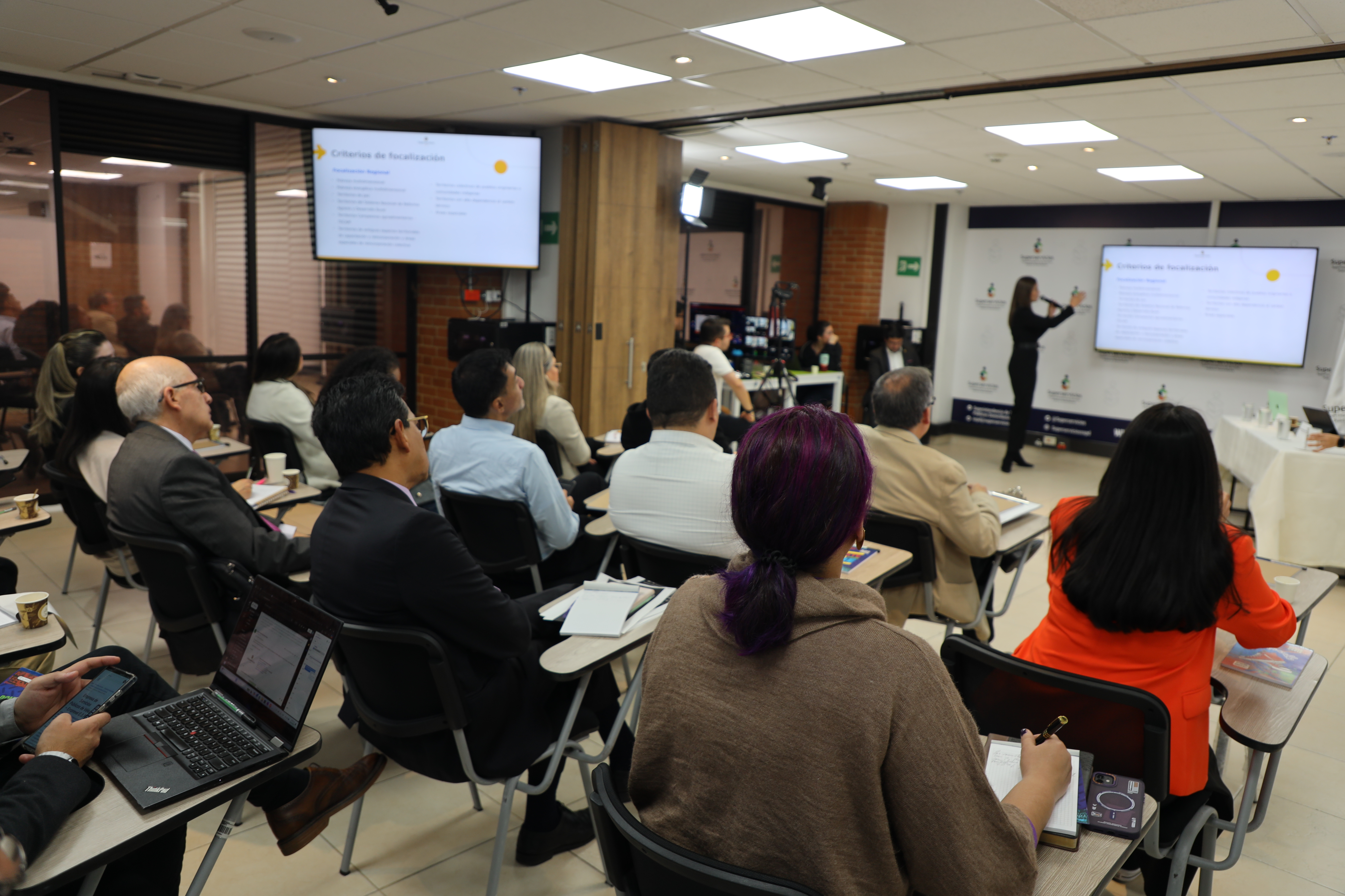 Personas sentadas en sillas, enfocadas en una pantalla, en un ambiente de discusión o aprendizaje.