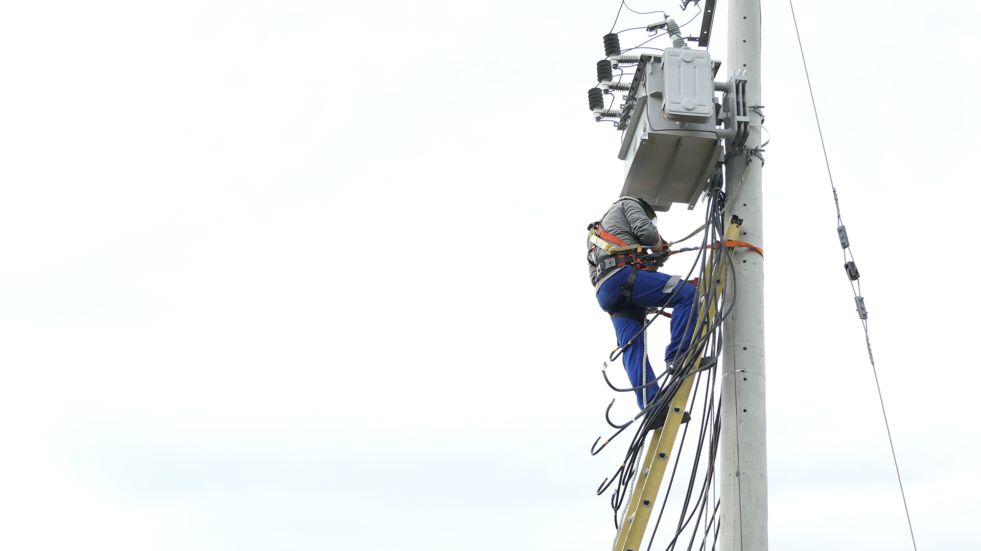 Un trabajador en un poste, ajustando una caja eléctrica, con equipo de seguridad visible en su vestimenta.